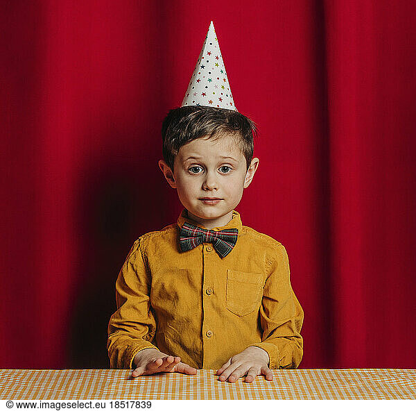 Boy in birthday hat sitting at table in front of red curtain