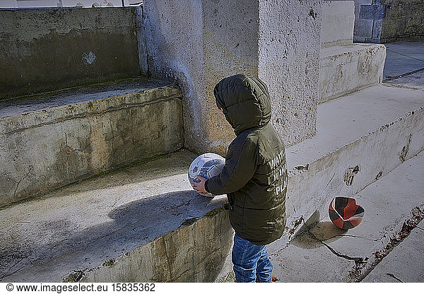Boy in blue trousers and green jacket playing ball on cement stairs.