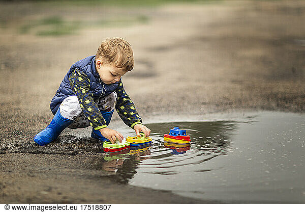 Boy in rain boots crouching in puddle and playing with ships