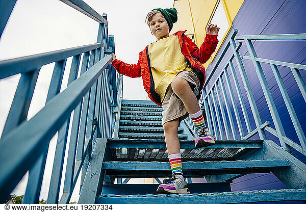 Boy in red jacket standing on one leg on metallic staircase by building