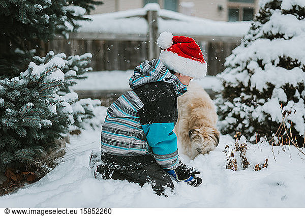 Boy in Santa hat playing in the snow with a dog in a yard.