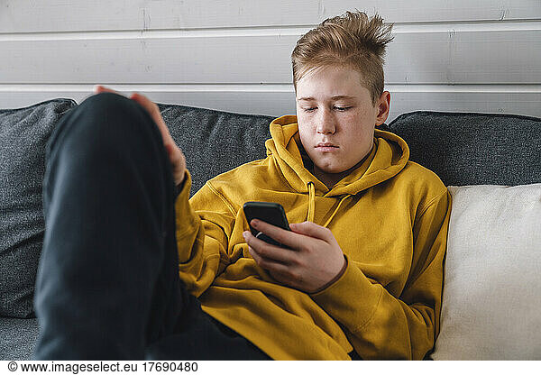 Boy in yellow hooded shirt using smart phone on sofa at home