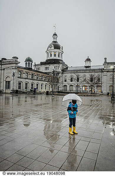 Boy in yellow rain boots holding umbrella in the rain in city square.