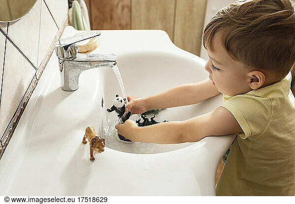 Boy in yellow t-shirt washing animal plastic toys in white sink