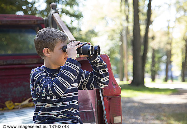 Boy leaning against pickup truck looking through binoculars in woods