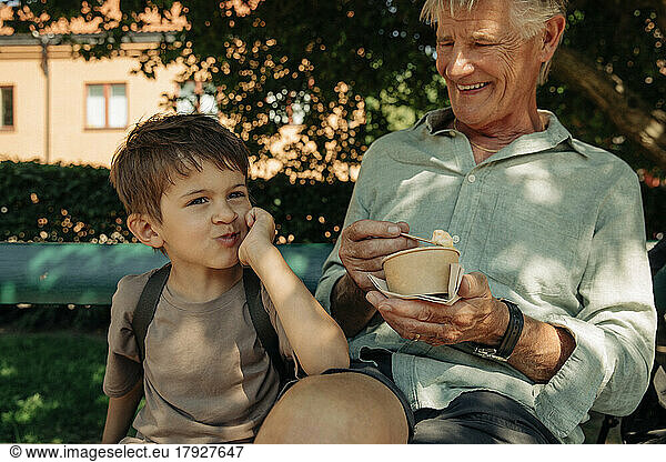 Boy leaning on elbow while sitting with grandfather holding ice cream cup at bench