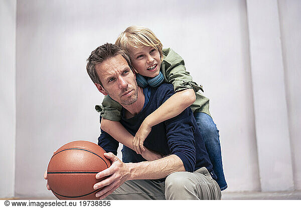 Boy leaning on father holding basketball in front of wall
