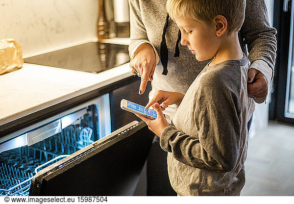 Boy learning from mother while using mobile app over dishwasher in kitchen at smart home