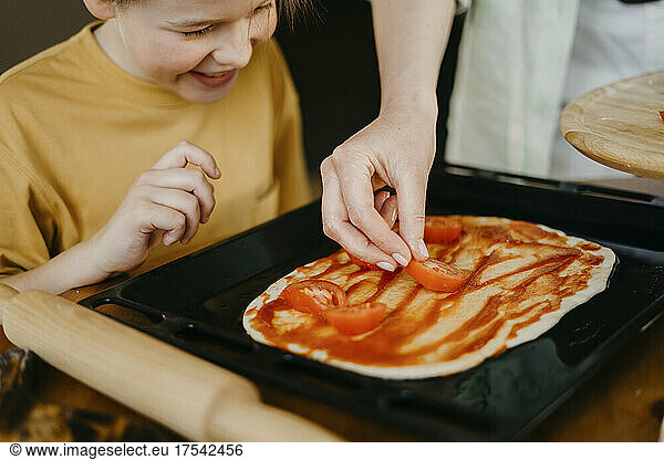 Boy looking at mother putting tomato slices on dough in tray