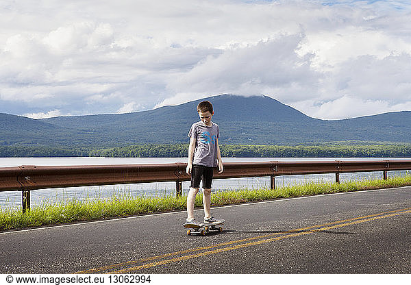 Boy looking down while standing on skateboard against cloudy sky