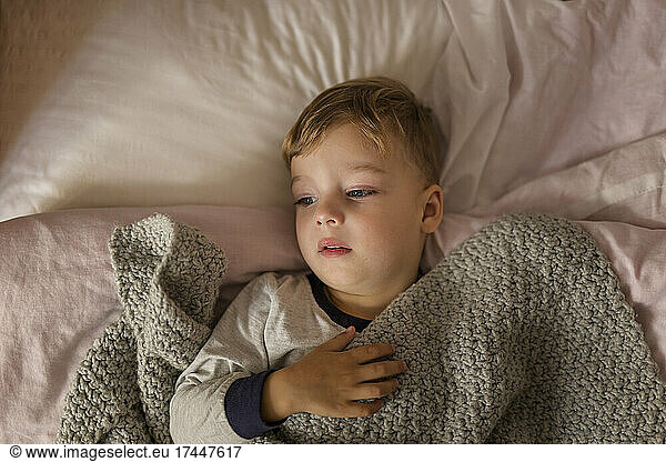 Boy lying on bed in pyjama being ill and covered with grey blanket