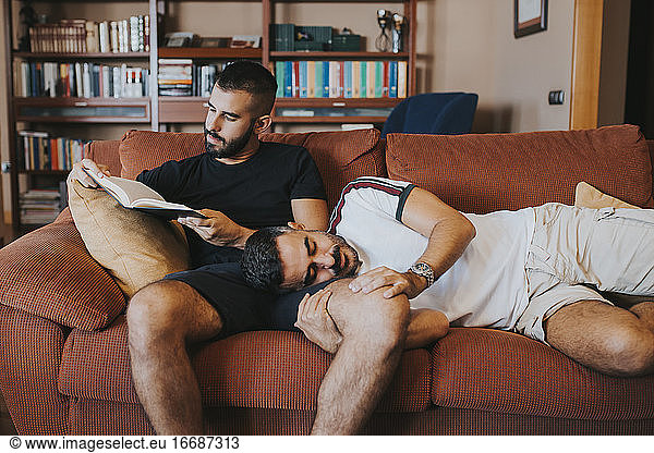 boy lying on his boyfriend while he reads a book on the sofa