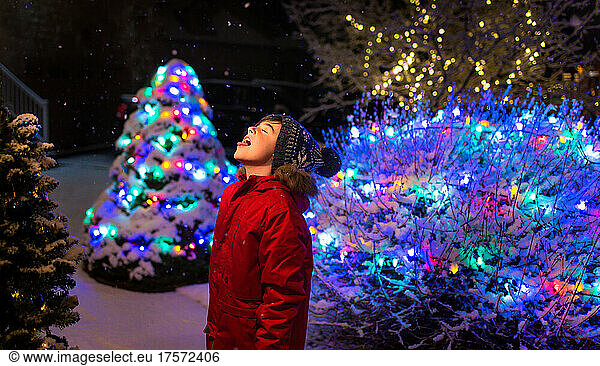 Boy outside at night in the snow with Christmas lights around him.