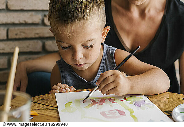 Boy painting on paper with mother in background at home