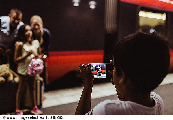 Boy photographing family on smart phone standing at railroad station platform