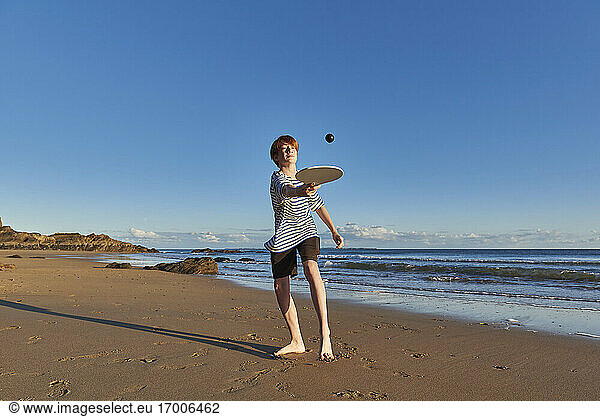 Boy playing with ball and racket while standing at beach