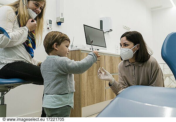 Boy playing with dental equipment while standing by dentist and mother wearing face mask in clinic