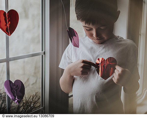 Boy playing with heart shape papers while standing by window at home