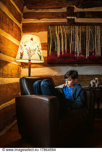 Boy reading a book alone in a chair in a cozy rustic log cabin.