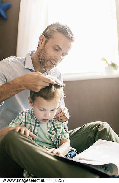 Boy reading book while father combing his hair at home