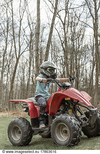 Boy riding a red four wheeler in grass with woods in background