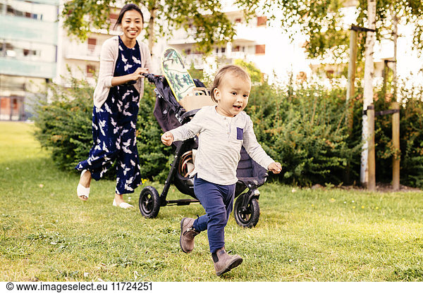 Boy (2-3) running on grass and mother walking with baby stroller in background