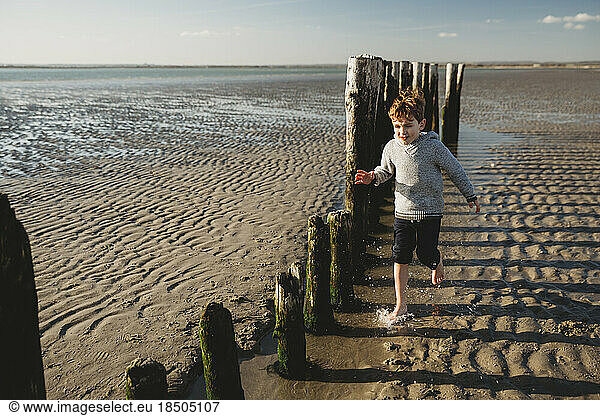 Boy running through water at beach against decaying breakwater pilings