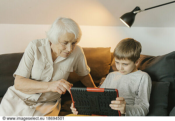 Boy sharing tablet PC with grandmother sitting on sofa at home