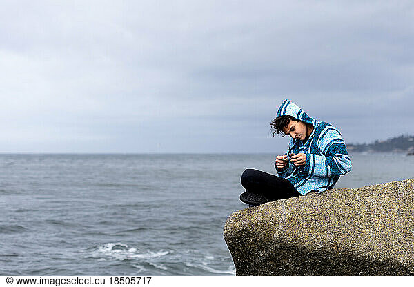 Boy Sitting On A Boulder Overlooking The Ocean Studies Rocks In Hand