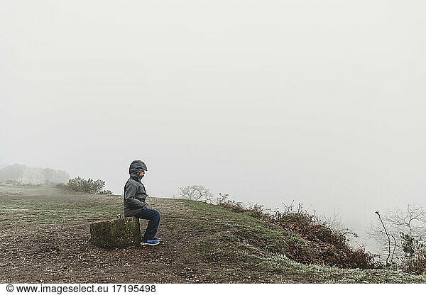 Boy sitting on concrete post in winter coat against foggy overcast sky