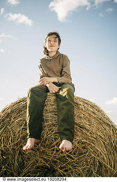 Boy sitting on haystack with sky in background
