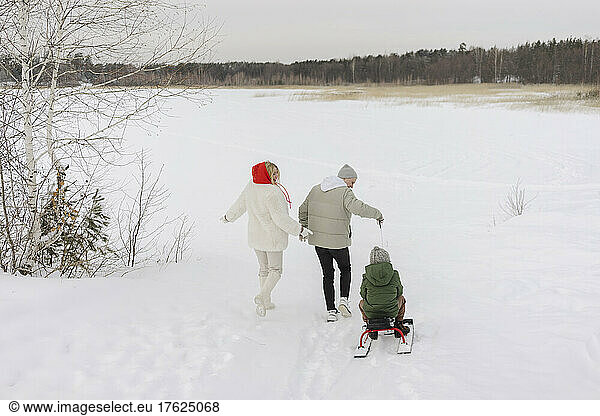 Boy sitting on toboggan pulled by father walking with woman on snow