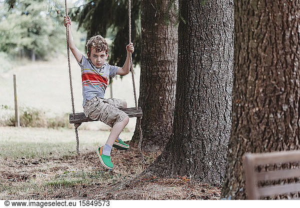 Boy sitting sideways on swing under pine trees