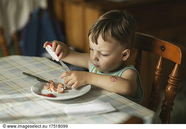 Boy sittyng by table and eating hot-dogs with fork