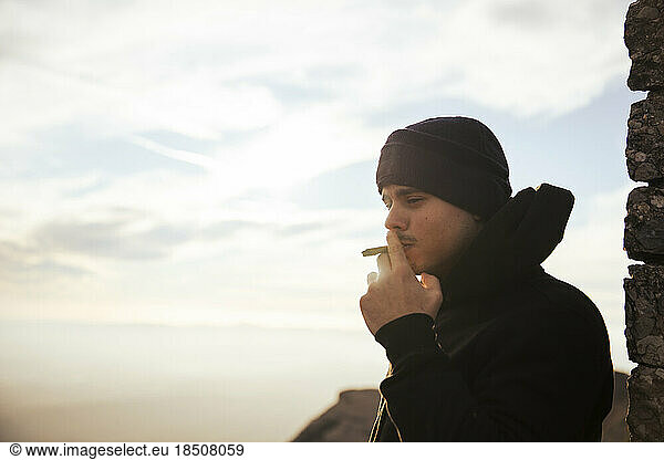 Boy smoking a marijuana joint on top of a mountain during sunset