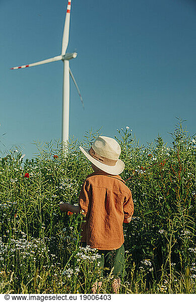 Boy standing in front of wind turbine and flowering plants at field