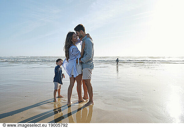 Boy standing with romantic parents dancing at beach