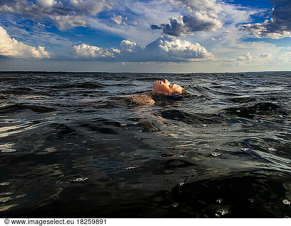 Boy swims in ocean water with white puffy cloud in blue sky summer
