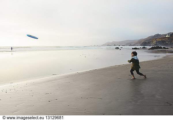 Boy throwing Frisbee while standing on shore at beach against sky