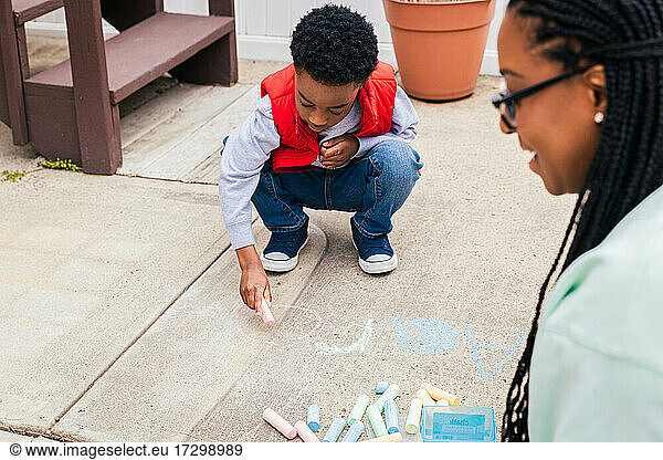Boy using chalk to draw on concrete while mother smiles and helps