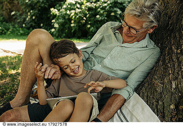 Boy using digital tablet while sitting with grandfather at park