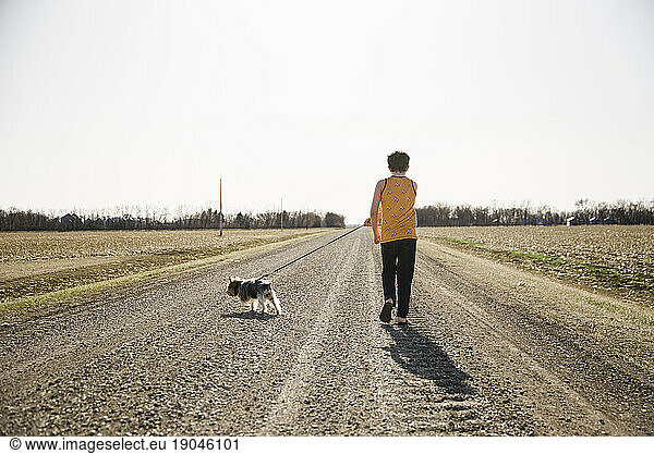 Boy walking his dog on a leash down a dirt road in the country.