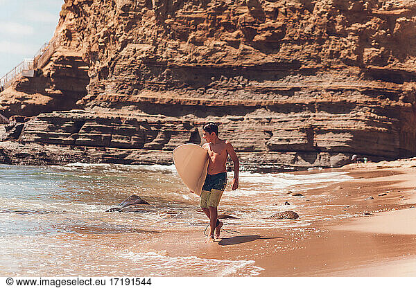 Boy walking on the beach carrying his surfboard - cliffs on the back.