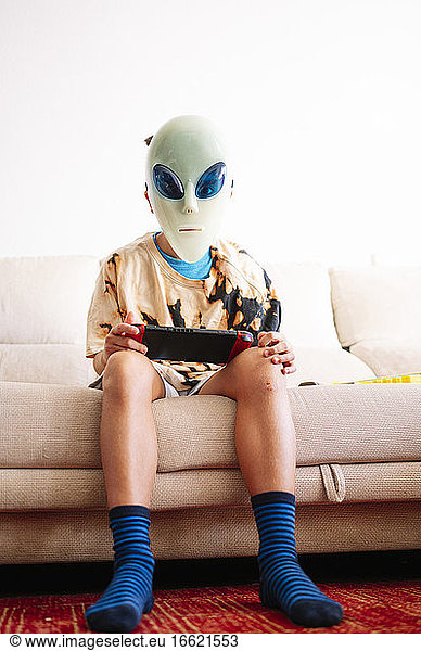 Boy wearing alien mask playing handheld video game while sitting on sofa at home