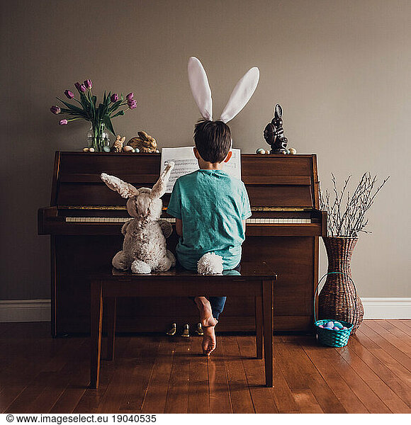 Boy wearing bunny ears playing piano with stuffed rabbit beside him.