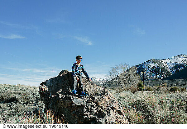Boy wearing sunglasses sits on huge rock snowy mountains in background