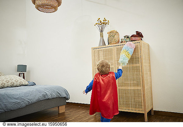 Boy wearing superman costume and cleaning with feather duster on a sideboard at home
