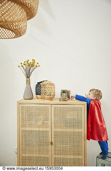 Boy wearing superman costume and reaching to wooden car on sideboard