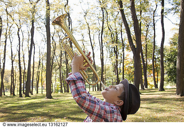 Boy wearing trilby playing trumpet in woods