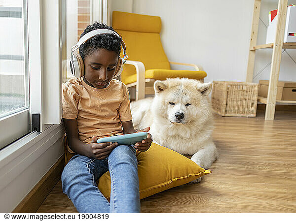 Boy wearing wireless headphones using mobile phone and sitting by dog at home
